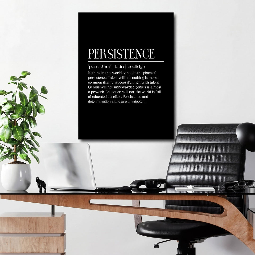 Motivational wall art with a black-and-white quote about persistence above a desk with a flower and open laptop