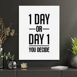 Motivational wall art with the black-and-white quote “1 DAY OR DAY 1 YOU DECIDE” in a modern office with plants and a clock.