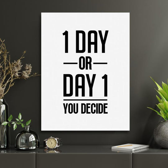 Motivational wall art with the black-and-white quote “1 DAY OR DAY 1 YOU DECIDE” in a modern office with plants and a clock.