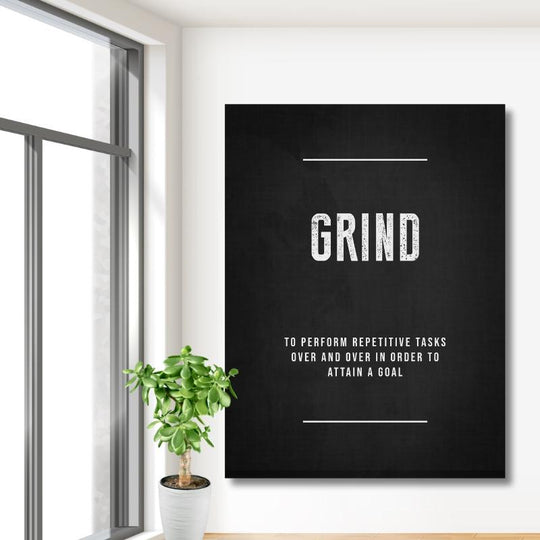 GRIND motivational canvas wall art in a modern office on a white wall near a large window and green plant.