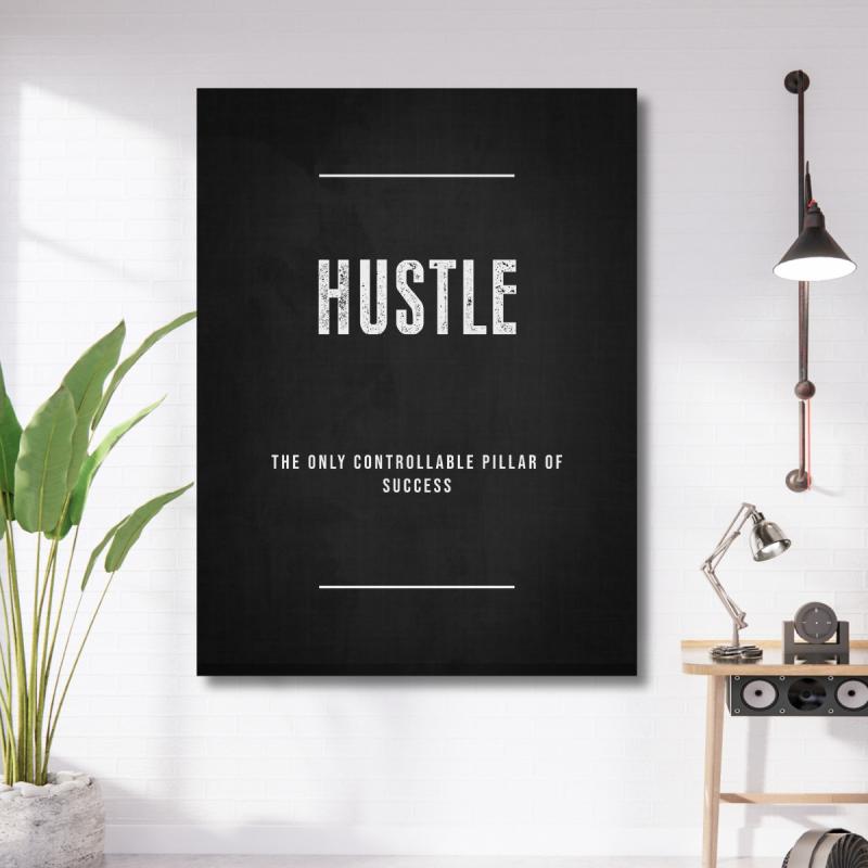 Black and white HUSTLE motivational canvas print in a minimalist office with a lamp and plant.